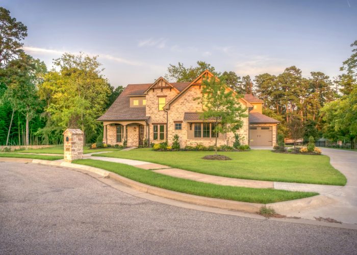 Elegant stone family home with manicured lawn, trees, and driveway on a sunny day.
